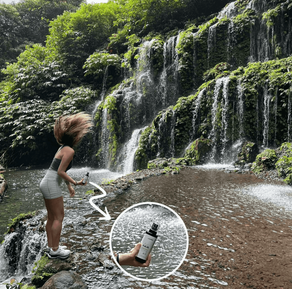 Femme tenant un spray feeds hair sur un rocher avec comme paysage une cascade d'eau